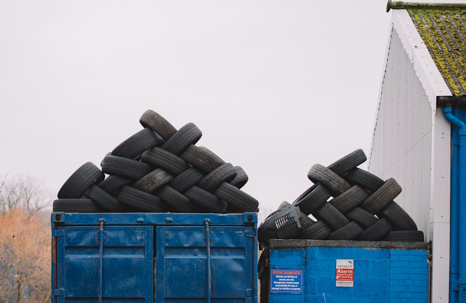 A photograph showing two large blue waste containers filled with stacked black rubber tires, positioned outdoors next to a white building with a moss-covered roof. The tires are piled in a crisscross pattern, with some leaning against the sides of the containers. The containers are situated on paved ground, with a cloudy sky overhead. In the background, a few leafless trees are visible, indicating a cold or winter season. The scene reflects waste management related to household or commercial tyre disposal, relevant to house removals and relocation services like those offered by Man with Van Queensbury, particularly when preparing for home relocation or furniture transport that requires clearing bulky items.