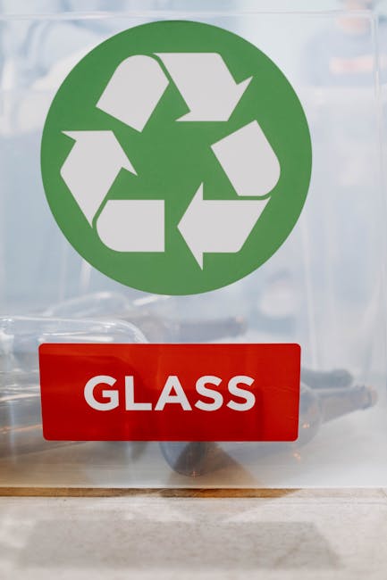 A clear glass container with a prominent red label reading 'GLASS', situated on a flat surface indoors. Behind the glass, a green recycling symbol consisting of three arrows forming a triangle is visible, indicating recyclable materials. The container appears to be used for collecting glass waste, with the environment suggesting an indoor space designated for waste sorting or collection as part of home relocation or packing and moving processes. The image is well-lit, and the background is slightly blurred to focus on the recycling symbol and glass container, aligning with services provided by Man with Van Queensbury for efficient furniture transport and waste disposal during house removals or moving logistics.