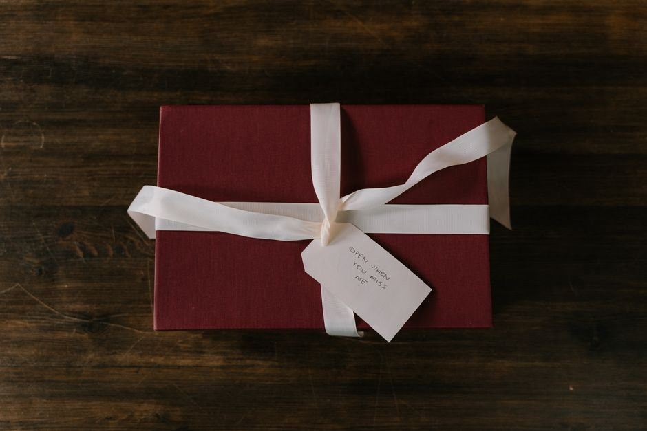 A rectangular gift box wrapped in dark red textured paper, tied with a white satin ribbon in a bow on top, rests on a dark wooden surface. Attached to the ribbon is a small white tag with the handwritten message 'Open when you miss me.' The background showcases the wooden surface, indicating an indoor setting, possibly during packing or home relocation preparations. The scene emphasizes careful packaging of personal belongings, often part of furniture transport or packing and moving processes involved in house removals, with natural lighting highlighting the textures of the wrapping materials and the wood grain.