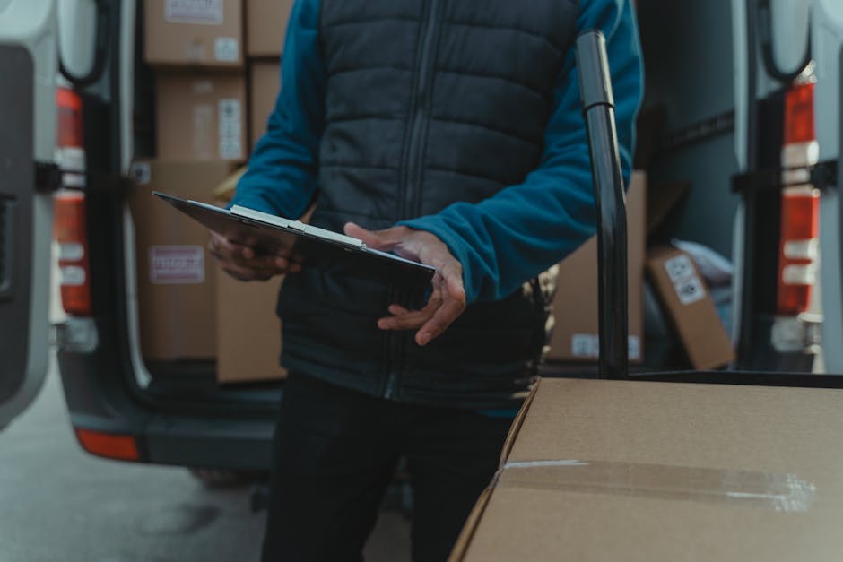 A person wearing a blue jacket and black pants is seen loading a cardboard box into the back of a van at a residential property during a house removal. The van's rear doors are open, revealing several