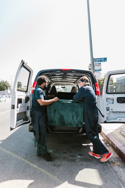A person wearing a blue jacket and black pants is seen loading a cardboard box into the back of a van at a residential property during a house removal. The van's rear doors are open, revealing several