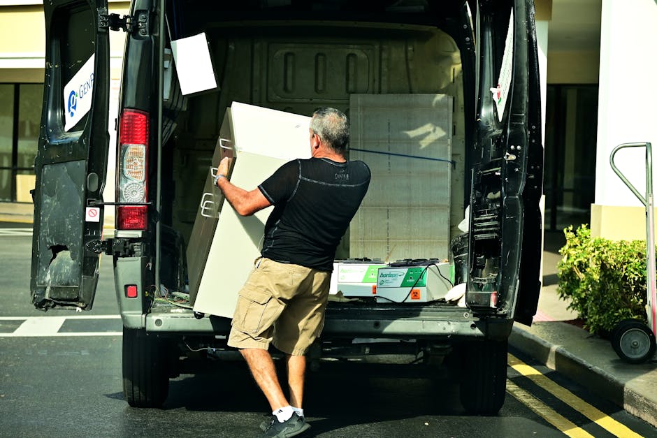 A man wearing a black t-shirt, beige shorts, and black shoes is seen loading a large white cabinet into the back of a black van parked on a street in Queensbury, within the HA3 postal district. The van's rear doors are wide open, revealing the interior filled with packing materials, cardboard boxes, and other furniture items awaiting transport during a home relocation. The man is using both hands to carefully position the cabinet inside the vehicle, which is part of the loading process for a house removal or furniture transport service. Nearby, on the pavement, a hand truck or dolly is visible, aiding the loading activities. In the background, a modern building with glass windows and some greenery can be seen, under bright daylight conditions, indicating an organized move by a professional removals company, such as Man with Van Queensbury, providing reliable loading and transport services.