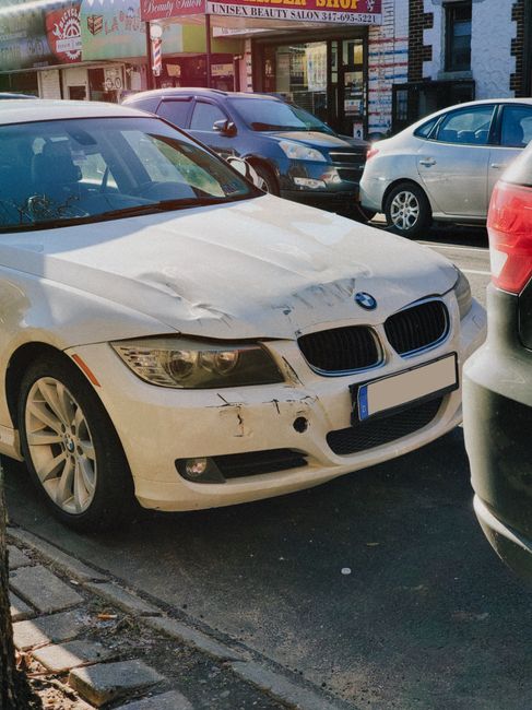 A white BMW sedan with visible front-end damage, including a crumpled hood, scratched bumper, and a missing front fog light, is parked on the street alongside other vehicles. Behind it, several cars are parked close together, including a dark grey van and a silver hatchback, in front of shopfronts with signs such as 'Beauty Salon' and 'Unisex'. The damaged car is positioned near the pavement, with its front facing the camera, and is part of a street scene showing parkers and storefronts in Queensbury. This image relates to house removals and furniture transport, highlighting the importance of careful vehicle and load management during home relocations and the potential for parking conflicts on busy streets. Man with Van Queensbury provides removals services that include navigating parking restrictions and managing logistics around street parking issues during moving processes.