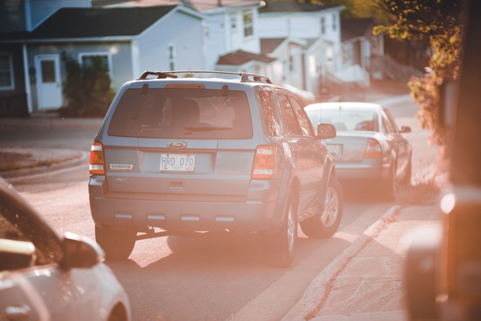 A photograph capturing a residential street scene during a house removal, showing two parked vehicles along the curb with a focus on the larger grey SUV positioned behind a smaller car. The vehicles are on a paved narrow street with a slight bend; the grey SUV, which appears to be a Ford, is situated close to a driveway of a house with white walls and a dark roof. In the background, there are several single-storey houses with white and blue facades, some with small front gardens, and a few trees with green foliage. The image is taken in warm, natural daylight, with sunlight casting a soft glow and slight lens flare present. The scene illustrates the logistics involved in packing and moving furniture or boxes, with the vehicles ready for transportation. Occasionally, Man with Van Queensbury's removals service manages such parking conflicts during home relocations, helping to facilitate smooth furniture transport and loading processes.