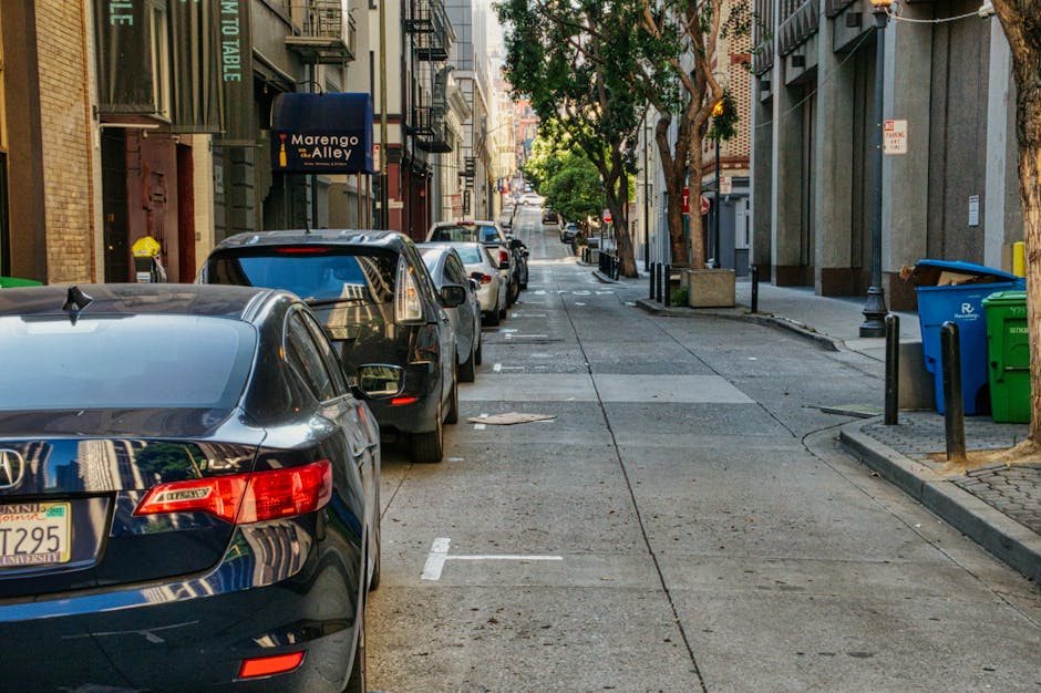 A white BMW sedan with visible front-end damage, including a crumpled hood, scratched bumper, and a missing front fog light, is parked on the street alongside other vehicles. Behind it, several cars are parked close together, including a dark grey van and a silver hatchback, in front of shopfronts with signs such as 'Beauty Salon' and 'Unisex'. The damaged car is positioned near the pavement, with its front facing the camera, and is part of a street scene showing parkers and storefronts in Queensbury. This image relates to house removals and furniture transport, highlighting the importance of careful vehicle and load management during home relocations and the potential for parking conflicts on busy streets. Man with Van Queensbury provides removals services that include navigating parking restrictions and managing logistics around street parking issues during moving processes.