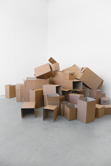 A large pile of cardboard moving boxes, various sizes, some with open flaps revealing empty interiors, positioned against a plain white wall in a spacious room with a smooth, light grey floor. The boxes are stacked haphazardly, with some leaning or partially collapsed, indicating a packing or home relocation process. Several of the boxes are wrapped with clear plastic or packing tape, and a few are placed in a random arrangement, suggesting either loading or unpacking. The scene captures the typical materials used in packing for furniture transport and moving services, with no furniture or appliances visible. This setup aligns with preparations often undertaken by professional removals companies like Man with Van Queensbury, who specialise in house removals, packing, and furniture transport within residential relocations.