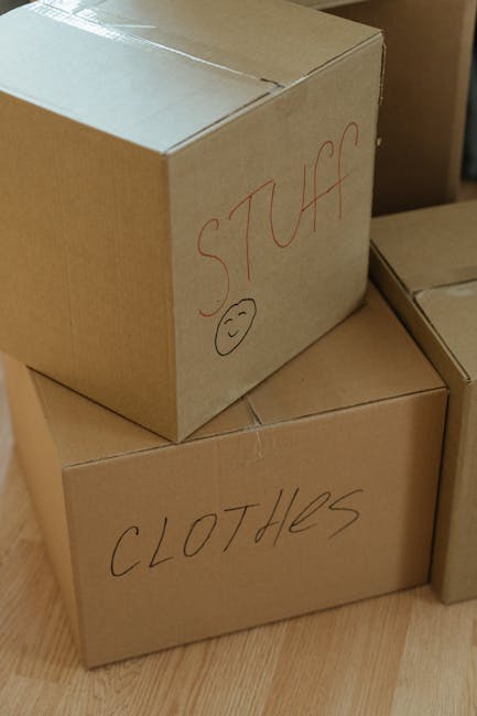 A large pile of cardboard moving boxes, various sizes, some with open flaps revealing empty interiors, positioned against a plain white wall in a spacious room with a smooth, light grey floor. The boxes are stacked haphazardly, with some leaning or partially collapsed, indicating a packing or home relocation process. Several of the boxes are wrapped with clear plastic or packing tape, and a few are placed in a random arrangement, suggesting either loading or unpacking. The scene captures the typical materials used in packing for furniture transport and moving services, with no furniture or appliances visible. This setup aligns with preparations often undertaken by professional removals companies like Man with Van Queensbury, who specialise in house removals, packing, and furniture transport within residential relocations.