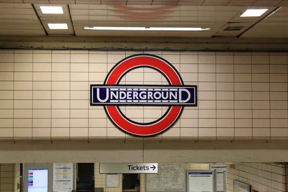 A large circular London Underground sign with a red outer ring, a blue bar across the center featuring the word 'UNDERGROUND' in white letters, mounted on a wall tiled in beige subway tiles within an underground station. Beneath the sign, there is a white sign with black text and an arrow pointing right that reads 'Tickets.' The station appears well-lit with ceiling panels and lights visible at the top of the image, but no people or moving objects are present in the scene, which provides a clear view of the signage and tiled interior typical of London tube stations.