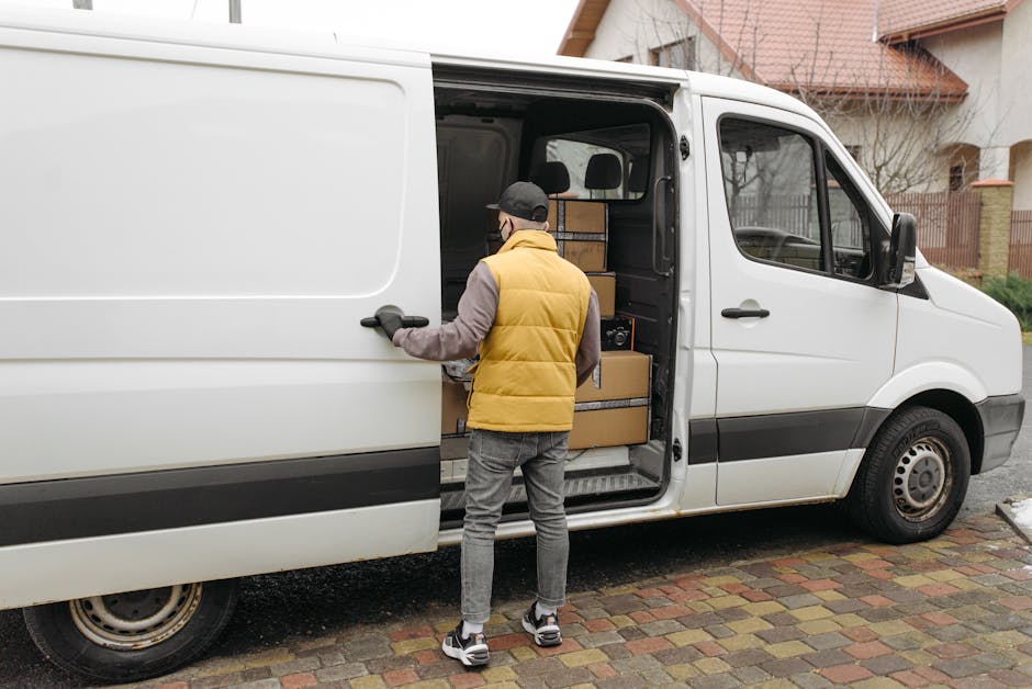 A man wearing a black cap, white t-shirt, and blue jeans stands beside a large moving van with the side door open, preparing for a home relocation. Inside the vehicle, visible equipment includes movin
