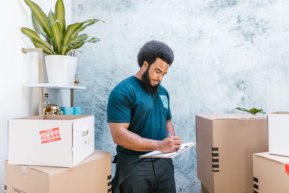 A convenience store employee with dark, curly hair and a beard is standing indoors among cardboard boxes, some labeled with red stickers indicating fragile items like glass. He is wearing a blue uniform shirt and is holding a clipboard while taking notes or checking a list. The boxes are stacked at different heights, with a large, leafy green plant on a white shelf in the background and a plain, textured light blue wall. The environment suggests packing and preparing items for a home relocation or moving service, with the employee documenting details as part of the packing and moving process. This scene is representative of a professional removals service such as Man with Van Queensbury, focused on furniture transport and logistics during an urgent same-day move.