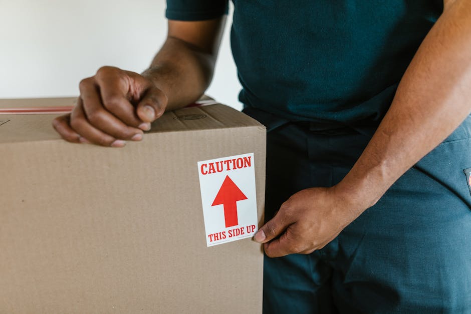 Close-up of a person wearing a dark green shirt and dark trousers, securely taping a large cardboard box sealed with packing tape. The box features a red and white caution label indicating 'Caution' and 'This Side Up' with an upward-pointing arrow, emphasizing proper handling during a home relocation. The individual is inside a house, near a staircase, preparing the box for transport or loading onto a van. The scene highlights the careful packing process involved in furniture transport and moving services. Behind the person, there is likely more packing material or additional boxes, contributing to an organized packing and moving setup. This image exemplifies the safety procedures and careful handling necessary during staircase moves in Queensbury, with [COMPANY_NAME] ensuring secure and professional furniture transport during house removals.