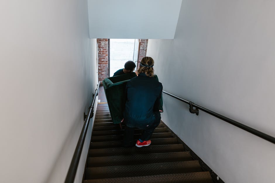 A person wearing a dark jacket, black pants, and red sneakers is crouching on a staircase while carrying a large, wrapped piece of furniture with a green cover during a home relocation. Ahead, two other individuals are visible, assisting with the move, with one wearing a dark jacket and the other with long hair tied back, both focused on lifting or guiding the furniture down the carpeted stairs. The staircase is narrow with dark treads and a black handrail on each side, leading down to a partly visible window at the bottom, which allows natural light into the corridor. Behind the movers, there is a brick wall with a partially visible window frame, indicating they are inside an older property, possibly during a furniture transport or packing and moving process. The image captures the careful handling involved in staircase moves, highlighting the importance of safe unloading procedures, an essential aspect of professional removals like those offered by Man with Van Queensbury.