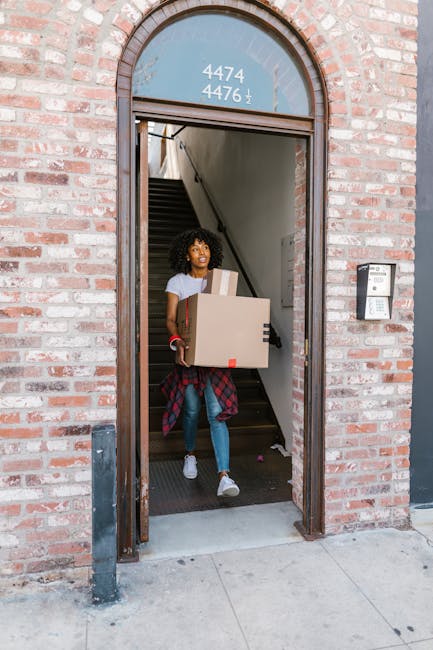 Close-up of a person wearing a dark green shirt and dark trousers, securely taping a large cardboard box sealed with packing tape. The box features a red and white caution label indicating 'Caution' and 'This Side Up' with an upward-pointing arrow, emphasizing proper handling during a home relocation. The individual is inside a house, near a staircase, preparing the box for transport or loading onto a van. The scene highlights the careful packing process involved in furniture transport and moving services. Behind the person, there is likely more packing material or additional boxes, contributing to an organized packing and moving setup. This image exemplifies the safety procedures and careful handling necessary during staircase moves in Queensbury, with [COMPANY_NAME] ensuring secure and professional furniture transport during house removals.